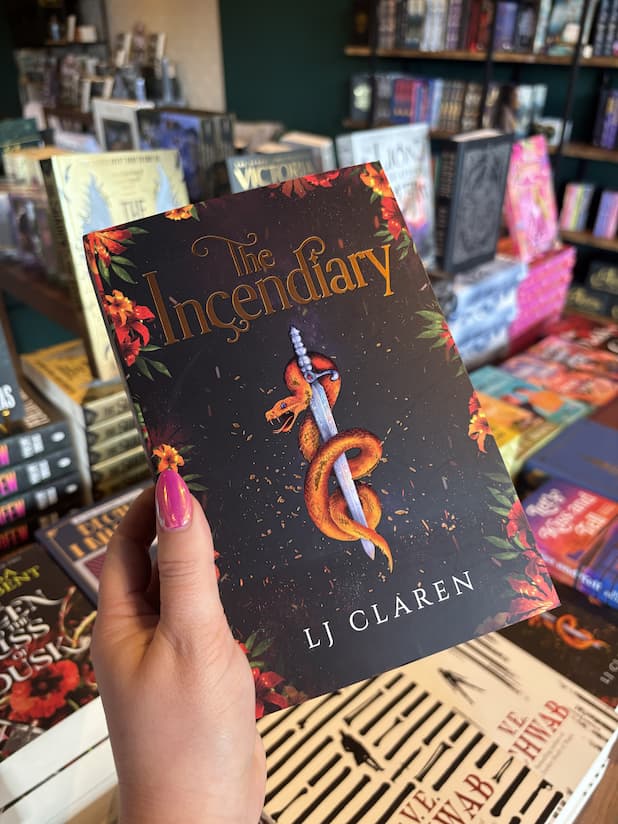 A hand with pink nails holding a cover for 'The Incendiary' by LJ Claren, featuring a golden-orange snake wrapped around a silver sword against a dark background, framed by red and orange flowers.