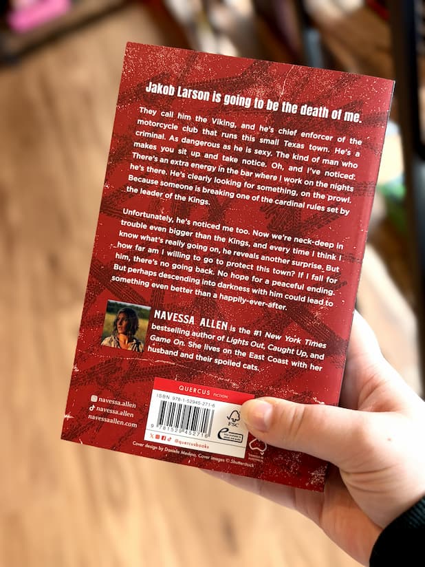 A hand holds the back of the book 'The Kings of Kearny' by Navessa Allen in front of a blurred bookshelf background. The cover is red and black with white text.