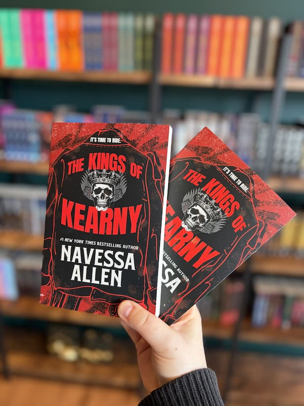 A hand holds two copies of the book 'The Kings of Kearny' by Navessa Allen in front of a blurred bookshelf background. The cover has a skull and crown design on a black and red background.