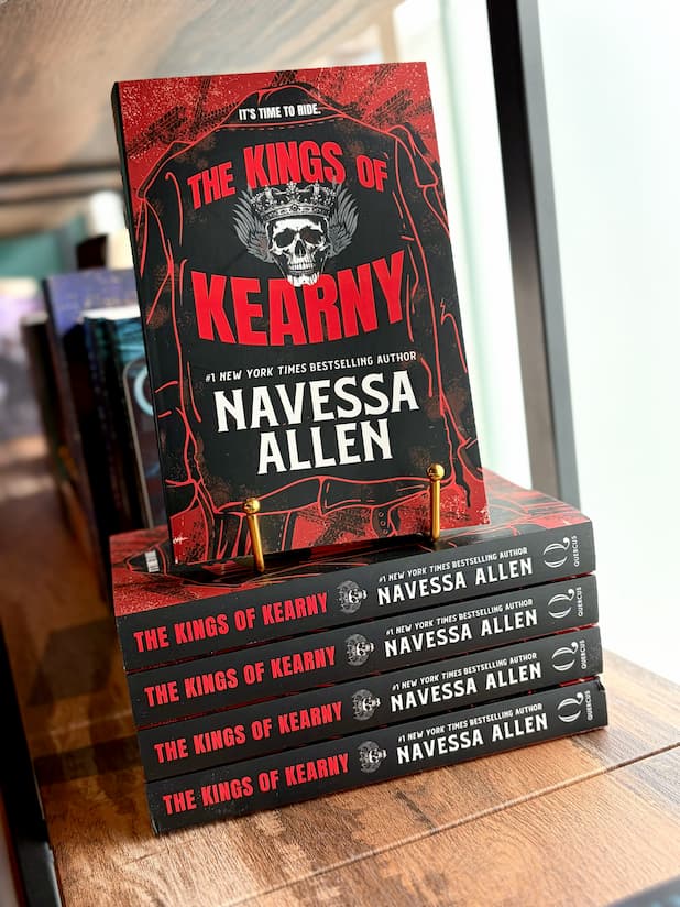 A stack of copies of the book 'The Kings of Kearny' by Navessa Allen, with one book standing upright to show the front cover, displayed on a wooden shelf. The cover has a skull and crown design on a black and red background.