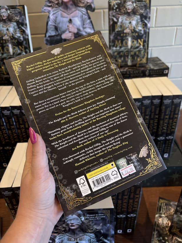A hand with pink nail polish holding the back of a paperback copy of 'The Knave and the Moon' by Rachel Gillig, featuring a dark background with moths, flowers, and white and yellow text. 