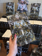 A hand with pink nail polish holding a paperback copy of 'The Knave and the Moon' by Rachel Gillig, features a woman in armour and a silken blindfold holding a longsword among smoke. The title is overlaid in gold with matching decorative corner borders. 