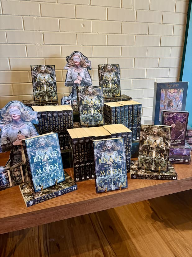 A display of several stacks of 'The Knave and the Moon' by Rachel Gillig. The cover features a woman in armour and a silken blindfold holding a longsword among smoke. The title is overlaid in gold with matching decorative corner borders. 