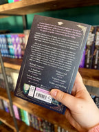 A hand holding the back of a copy of 'The Knight and the Moth' by Rachel Gillig, featuring a dark textured background with white and pink text.