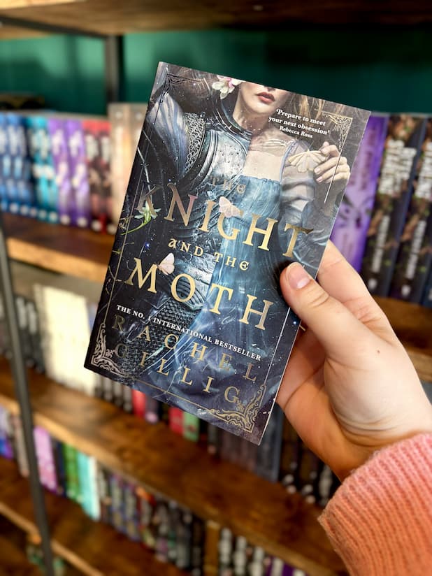 A hand holding a copy of 'The Knight and the Moth' by Rachel Gillig, featuring a woman lying in a body of water. Half of her is in a light blue dress and the other half is in a suit of armour. There are white moths scattered about and the title is in large white text.