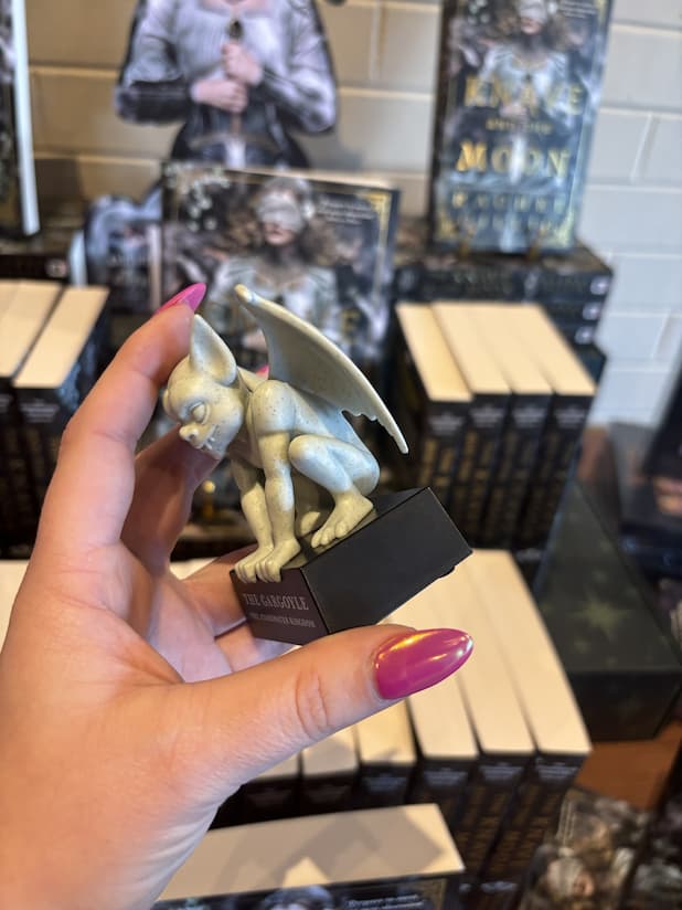 A hand with pink nail polish holding a gargoyle figurine to the side. It sits upon a platform that reads 'The Gargoyle' in large text and 'The Stonewater Kingdom' in smaller text under that.