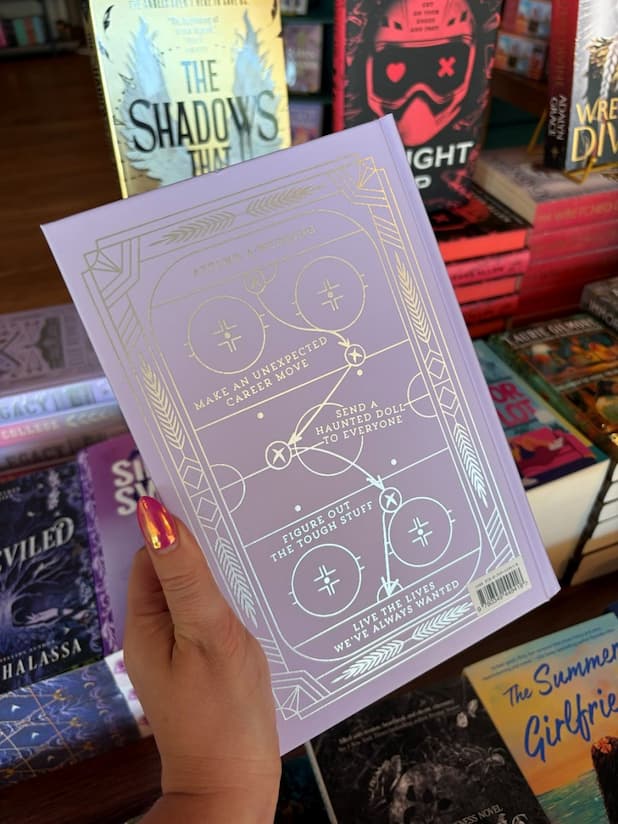 A hand with pink nail polish holds the back of a hardcover special edition of 'The Legacy' by Elle Kennedy, featuring a pastel purple with a silver hockey play drawn out with a silver border.