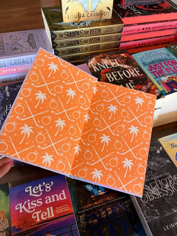 A hand with pink nail polish holds open a hardcover special edition of 'The Legacy' by Elle Kennedy, featuring orange pages with white illustrations of hockey sticks and palm trees.