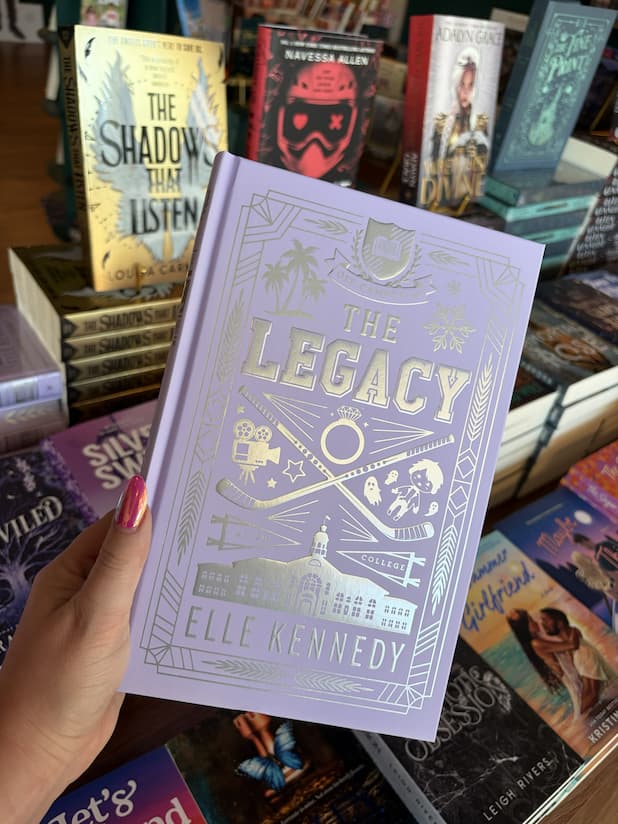 A hand with pink nail polish holds a hardcover special edition of 'The Legacy' by Elle Kennedy, featuring a pastel purple with silver text and hockey illustrations.