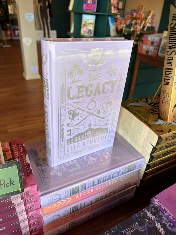 A hardcover copy of the book 'The Legacy' by Elle Kennedy, standing upright on a stack of other copies in a bookstore setting. The cover features a pastel purple with silver text and hockey illustrations.