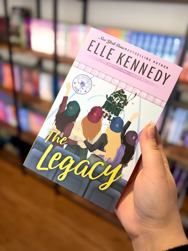 A hand holding an alt paperback copy of 'The Legacy' by Elle Kennedy, featuring a glossy illustration of four women waving at men on an ice rink.