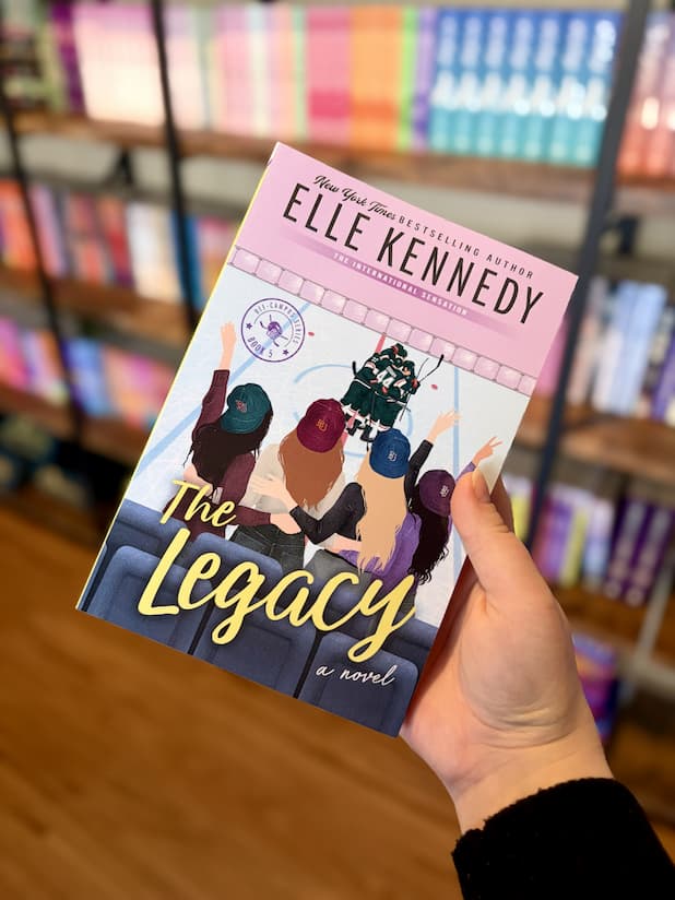 A hand holding a paperback copy of 'The Legacy' by Elle Kennedy, featuring an illustration of four women waving at men on an ice rink.