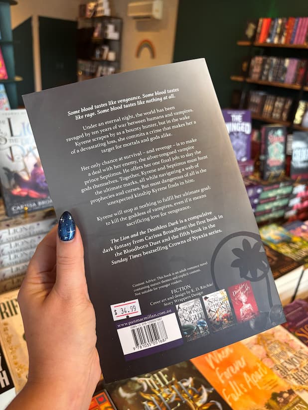 A hand with blue glitter nail polish holds a book titled 'The Lion and the Deathless Dark' by Carissa Broadbent, showing the back cover with synopsis text and publisher logos, with a blurred bookstore background.