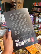 A hand with blue glitter nail polish holds a book titled 'The Lion and the Deathless Dark' by Carissa Broadbent, showing the back cover with synopsis text and publisher logos, with a blurred bookstore background.