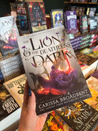 A hand holds a copy of the book 'The Lion & the Deathless Dark' by Carissa Broadbent in a book store, with other book spines visible in the background.