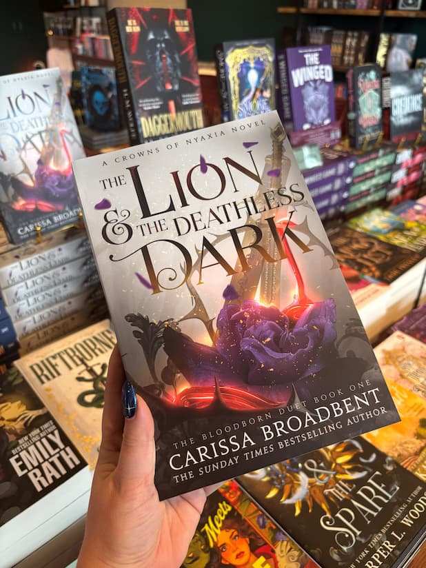 A hand holds a copy of the book 'The Lion & the Deathless Dark' by Carissa Broadbent in a book store, with other book spines visible in the background.
