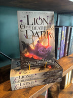 A copy of the book 'The Lion & the Deathless Dark' by Carissa Broadbent is displayed upright on top of two matching copies on a wooden bookshelf, with other books visible in the background.