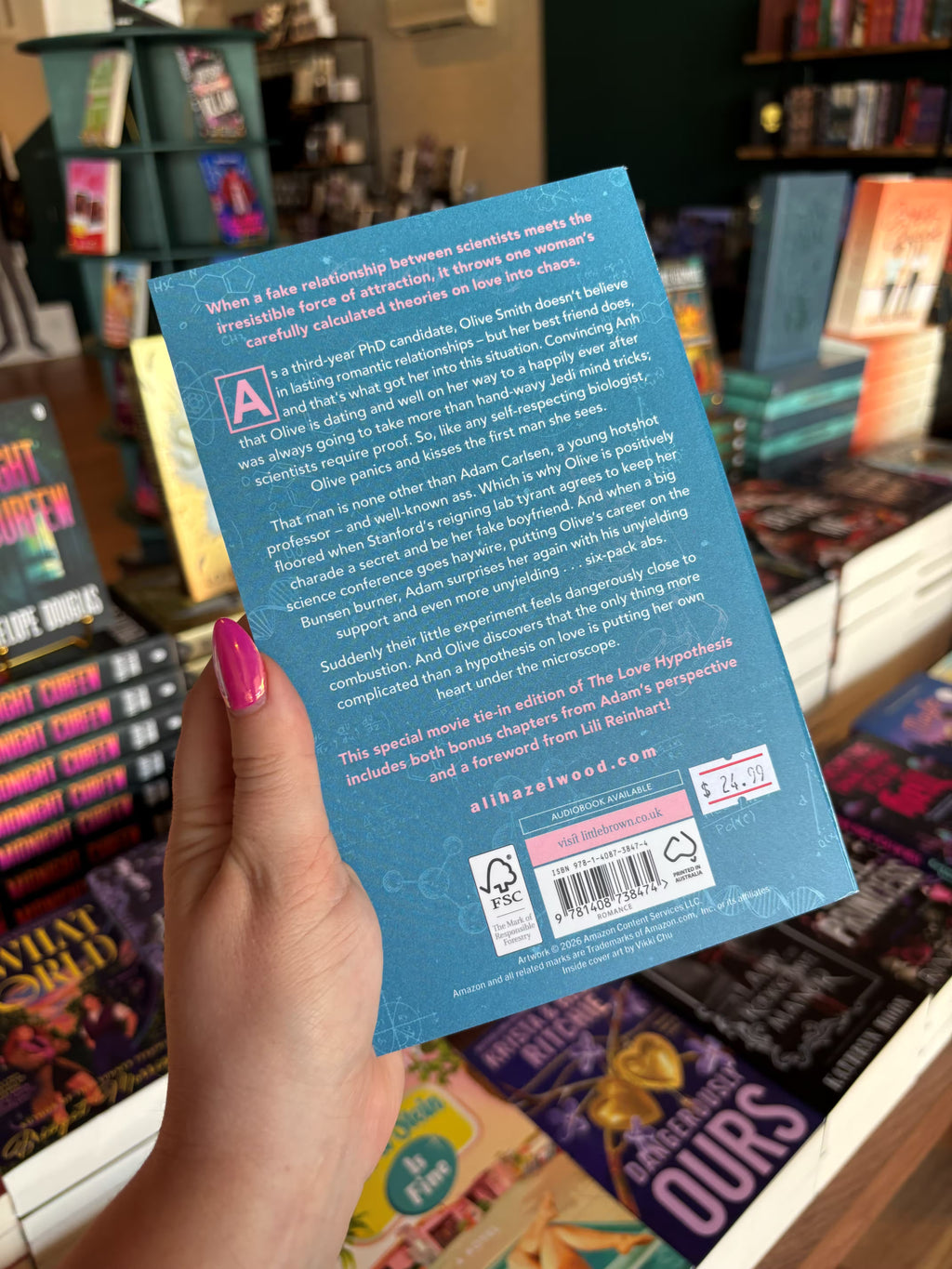 A hand with pink nail polish holds the back cover for 'The Love Hypothesis' by Ali Hazelwood in a bookshop with other books blurry in the background. The cover features a blue background with scientific diagrams and white text for the blurb.