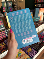 A hand with pink nail polish holds the back cover for 'The Love Hypothesis' by Ali Hazelwood in a bookshop with other books blurry in the background. The cover features a blue background with scientific diagrams and white text for the blurb.