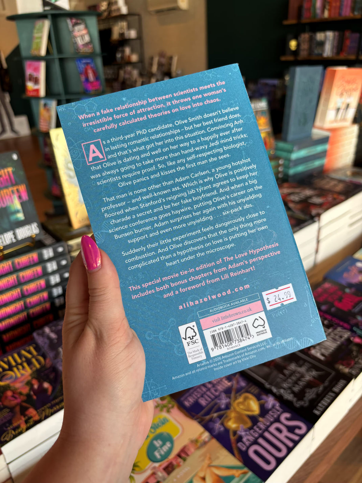 A hand with pink nail polish holds the back cover for 'The Love Hypothesis' by Ali Hazelwood in a bookshop with other books blurry in the background. The cover features a blue background with scientific diagrams and white text for the blurb.