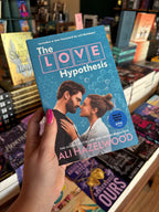 A hand with pink nail polish holds the book cover for 'The Love Hypothesis' by Ali Hazelwood in a bookshop with other books blurry in the background. The cover features a man and woman facing each other on a blue background with periodic-table-style lettering for 'LOVE' and scientific diagrams.