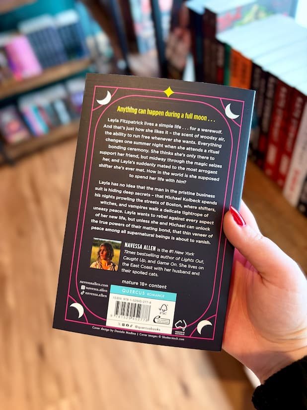 A hand holding the back cover of 'The Lunatics' by Navessa Allen, with a blurred background of other books on a wooden surface.