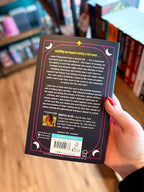 A hand holding the back cover of 'The Lunatics' by Navessa Allen, with a blurred background of other books on a wooden surface.