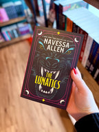 A hand with red nail polish holds a paperback book titled 'The Lunatics' by New York Times Bestselling Author Navessa Allen. The book cover features a black background with a blue graphic of a wolf with the title between his teeth
