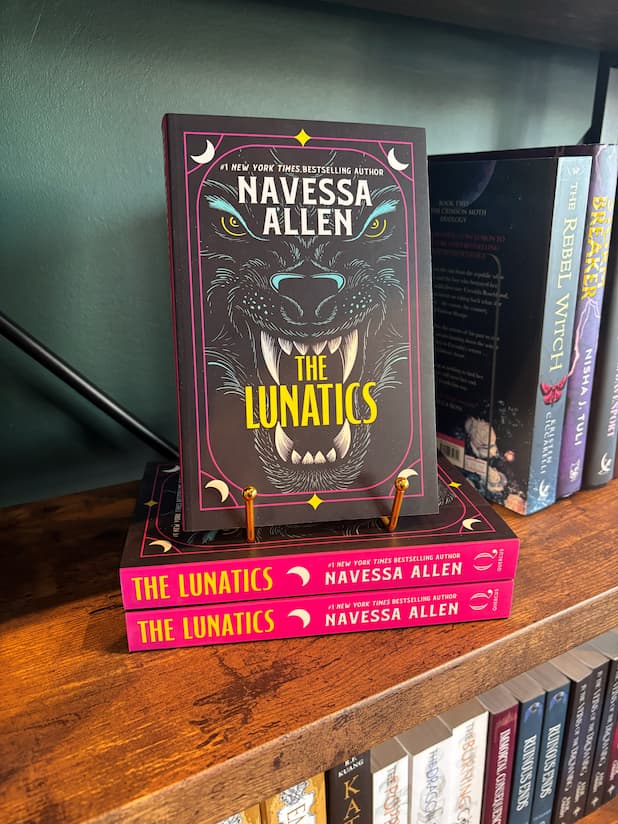 A copy of the book 'The Lunatics' by New York Times Bestselling Author Navessa Allen displayed on a wooden bookshelf, standing upright on a small gold book stand next to other copies of the book.