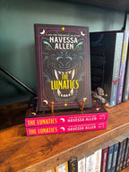 A copy of the book 'The Lunatics' by New York Times Bestselling Author Navessa Allen displayed on a wooden bookshelf, standing upright on a small gold book stand next to other copies of the book.