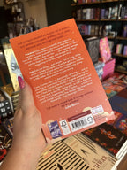 A hand with pink nail polish holding the back of a copy of 'The Magic We Made' by Meg Cabot with an orange background and white and brown text.