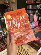 A hand with pink nail polish holding a copy of 'The Magic We Made' by Meg Cabot with a graphic illustration of a man and a woman in a vintage car, house, and sunset all on an orange toned background.