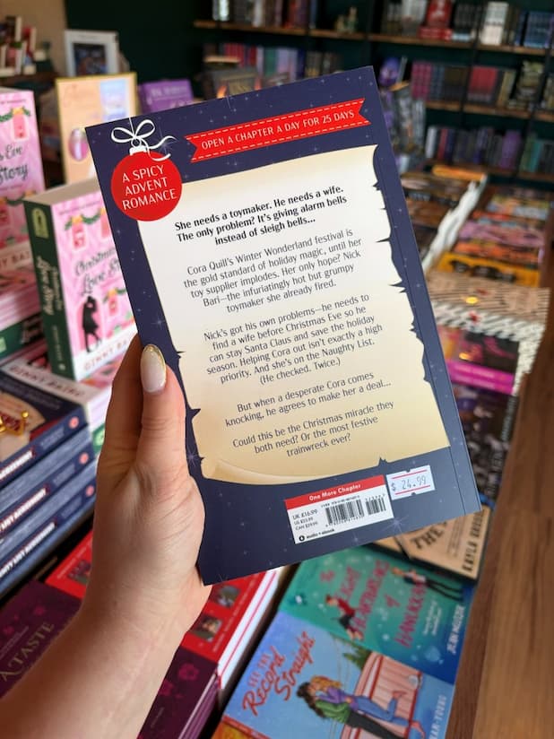 A hand with clear nail polish holding the back of a copy of 'The Naughty List Agreement' by Katherine Garbera. featuring a list with the blurb, a bauble and a blue starry background. 