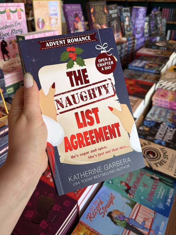 A hand with clear nail polish holding a copy of 'The Naughty List Agreement' by Katherine Garbera, featuring a woman with red nails in a santa outfit holding a list with the title with holly, a bauble and a blue starry background. 