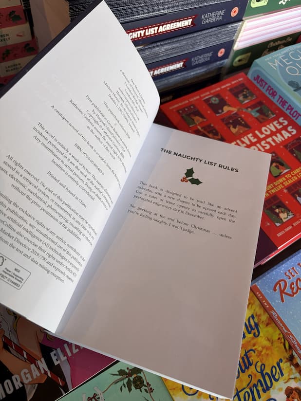 A hand with clear nail polish holding open a copy of 'The Naughty List Agreement' by Katherine Garbera, featuring interior white pages with the rules of the book.