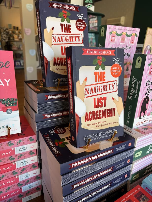 Two stacks of 'The Naughty List Agreement' by Katherine Garbera, featuring a woman with red nails in a santa outfit holding a list with the title with holly, a bauble and a blue starry background. 