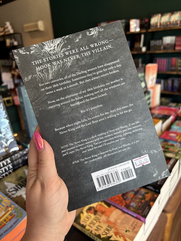 A hand with pink nail polish holding the back of a copy of 'The Never King' with  white text on a dark textured background. 