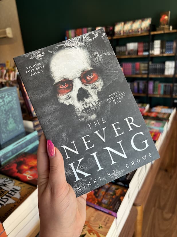 A hand with pink nail polish holding a copy of 'The Never King' with a skull and red eyes on a dark background.