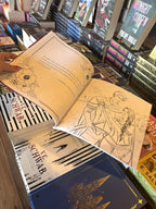 A hand with pink nail polish holding an open copy of 'The Official Blood and Ash Coloring Book' by Jennifer L. Armentrout, featuring a quote on the left page and line art of a couple in armour on the right page.