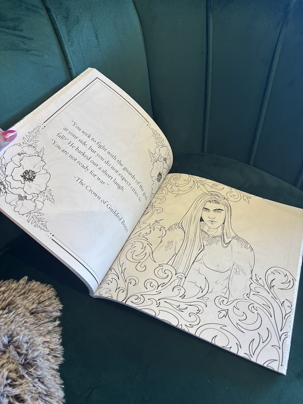 A hand with pink nail polish holding the back of a copy of 'The Official Blood and Ash Coloring Book' by Jennifer L. Armentrout, featuring a quote on the left page and a shirtless man with long hair on the right page.