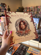 A hand with pink nail polish holding a copy of 'The Official Blood and Ash Coloring Book' by Jennifer L. Armentrout, featuring a cream background with red detailing as the border and a woman's portrait in the centre.  