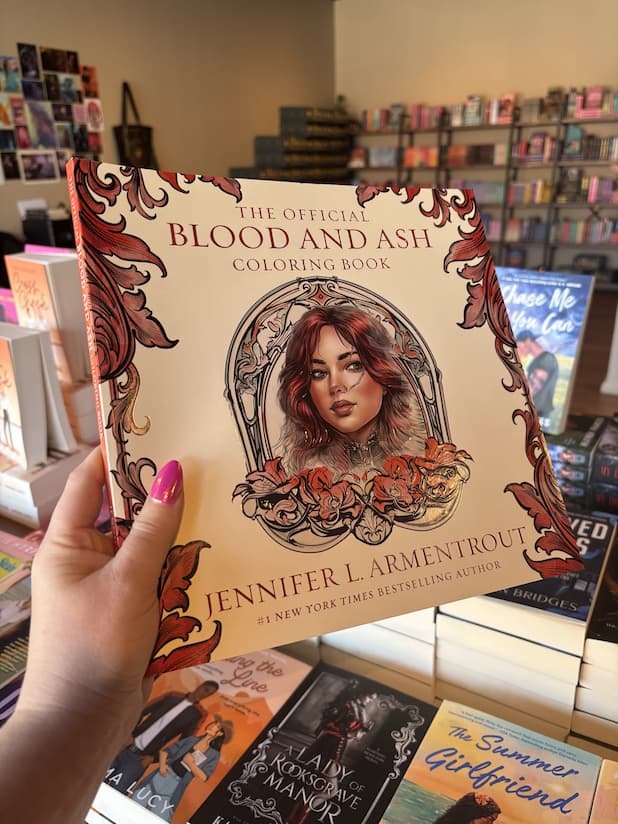 A hand with pink nail polish holding a copy of 'The Official Blood and Ash Coloring Book' by Jennifer L. Armentrout, featuring a cream background with red detailing as the border and a woman's portrait in the centre.  