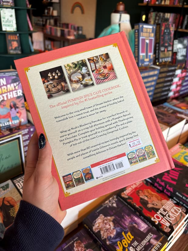 A hand holding the back cover of 'The Official Pumpkin Spice Cafe Cookbook' from the Dream Harbour Series, with a background of other books on a wooden surface.