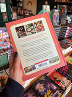 A hand holding the back cover of 'The Official Pumpkin Spice Cafe Cookbook' from the Dream Harbour Series, with a background of other books on a wooden surface.