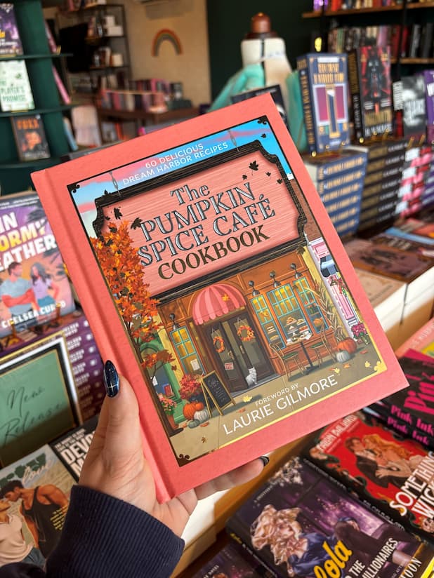 A hand with red blue starry nail polish holds a copy of 'The Official Pumpkin Spice Café Cookbook' by Heather Thomas and Laurie Gilmore from the Dream Harbour Series. The book cover features a orange border around an illustrated picture of a cafe.