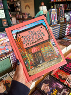 A hand with red blue starry nail polish holds a copy of 'The Official Pumpkin Spice Café Cookbook' by Heather Thomas and Laurie Gilmore from the Dream Harbour Series. The book cover features a orange border around an illustrated picture of a cafe.