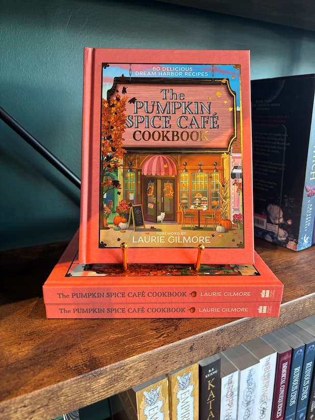 A copy of the book 'The Official Pumpkin Spice Café Cookbook' by Heather Thomas and Laurie Gilmore displayed on a wooden bookshelf, standing upright on a small gold book stand next to other copies of the book.