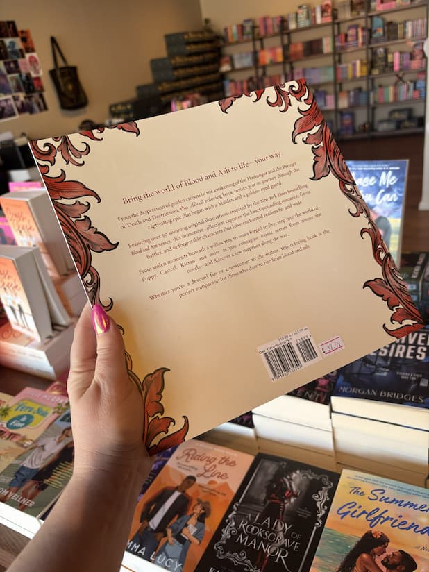 A hand with pink nail polish holding the back of a copy of 'The Official Blood and Ash Coloring Book' by Jennifer L. Armentrout, featuring a cream background with red detailing and red text.
