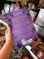 A hand with blue nail polish holds a purple book cover copy of 'The Poison Daughter' by Sheila Masterson showing the back cover in a bookstore with other books visible in the background.