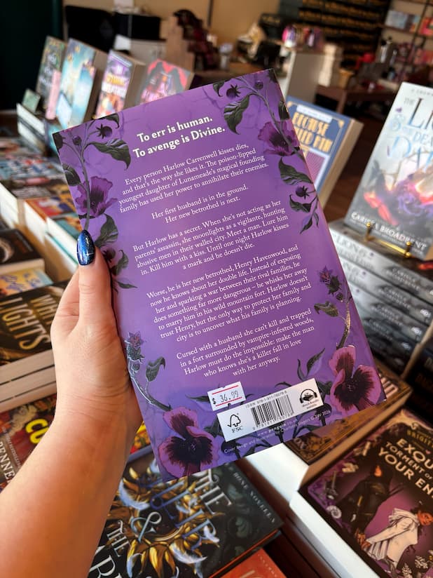 A hand with blue nail polish holds a purple book cover copy of 'The Poison Daughter' by Sheila Masterson showing the back cover in a bookstore with other books visible in the background.